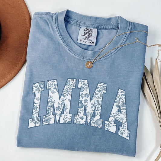 Folded blue t-shirt with 'IMMA' design on a white surface with a hat and dried leaves.