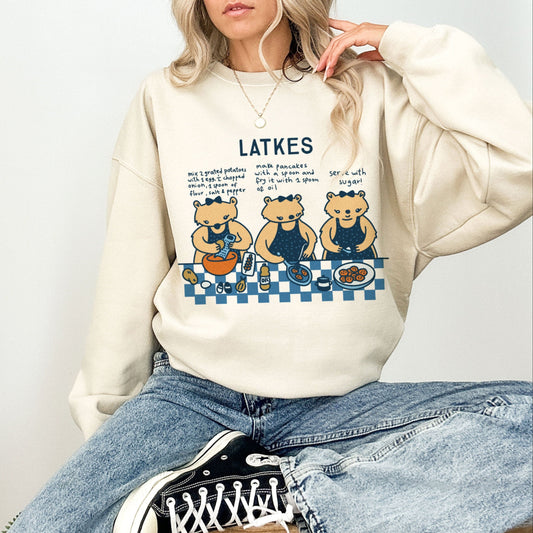 A person wearing a beige sweatshirt with a graphic of four cartoon bears around a table eating latkes, with the word 'LATKES' above.
